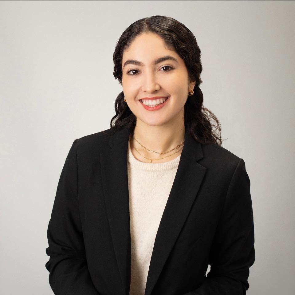 A young woman with dark wavy hair smiling, wearing a black blazer, white shirt, and gold jewelry, standing against a light-colored background.