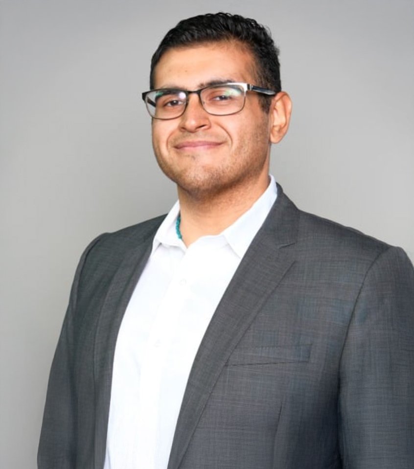 Professional man in a suit with glasses, smiling, against a plain gray background.