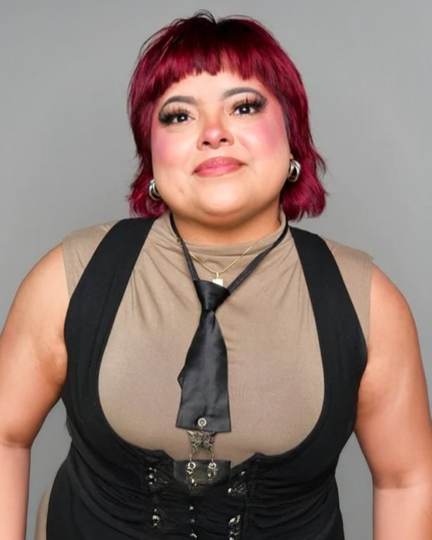 A woman with short, vibrant red hair, fair skin, and makeup with bold eyeliner and blush, wearing a beige sleeveless top, a black vest, a black tie, and jewelry including hoop earrings and layered necklaces, against a gray background.