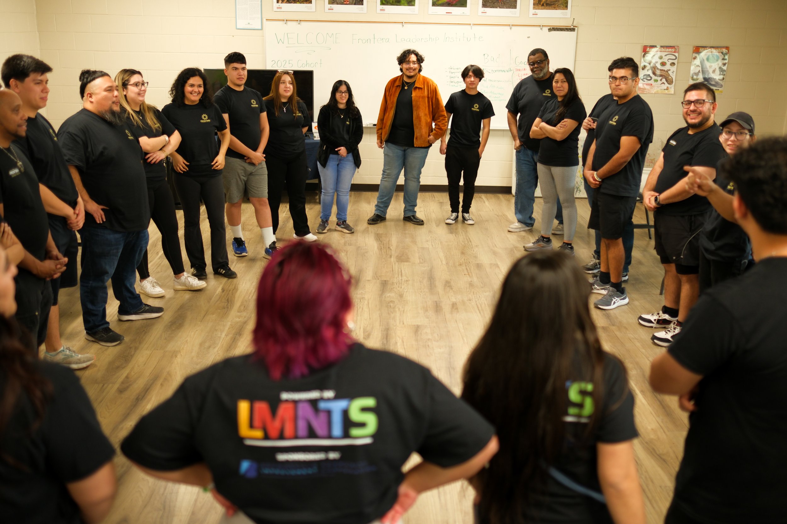 Group of diverse people standing in a circle in a classroom, participating in a team-building activity, with some seated in the foreground.