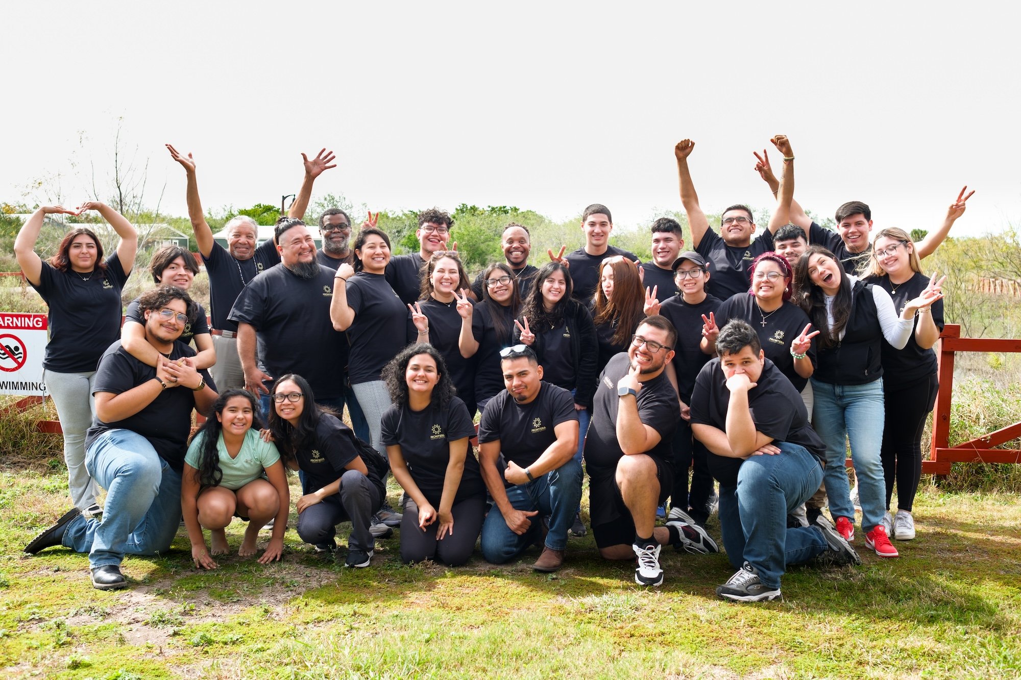 Group of smiling diverse young adults and middle-aged people posing outdoors, some making peace signs and other gestures, on a grassy area with trees in the background.
