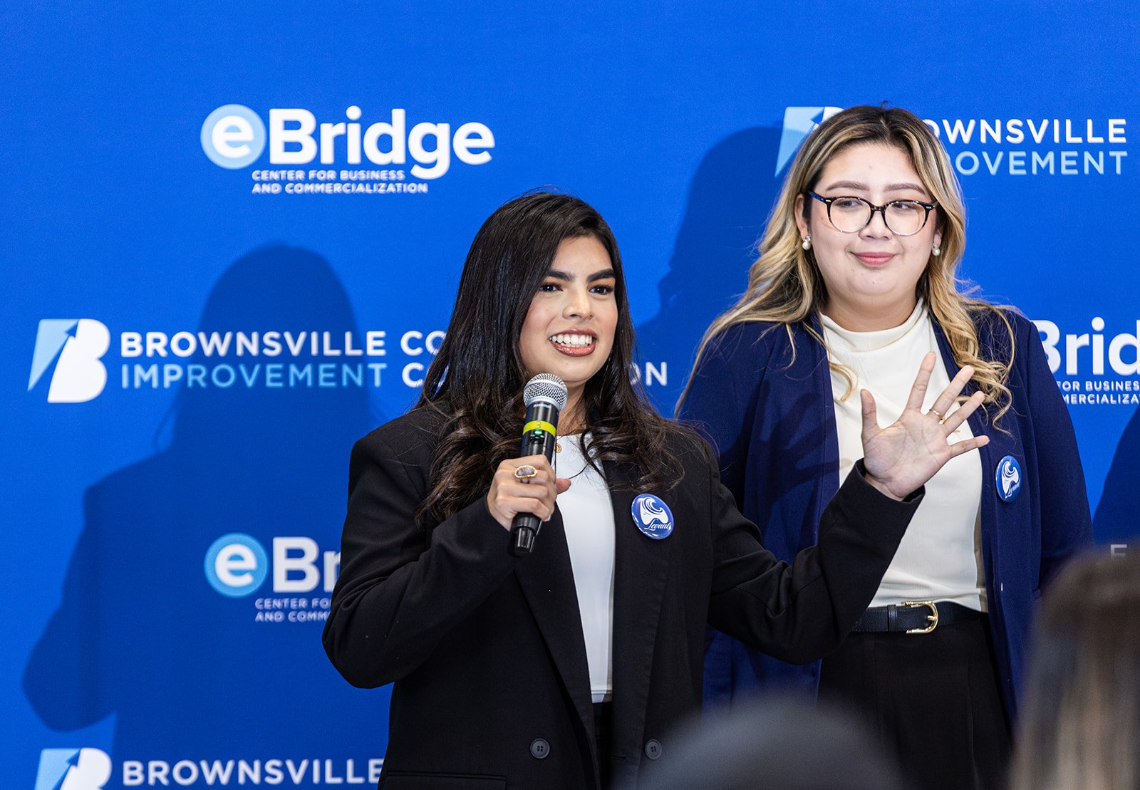Two women speaking at an event with a blue background that displays the texts 'eBridge', 'Center for Business and Commercialization', 'Brownsville Improvement', and 'Brownsville'. The woman on the left is holding a microphone and wearing a black blazer, while the woman on the right is waving and wearing glasses and a blue blazer.