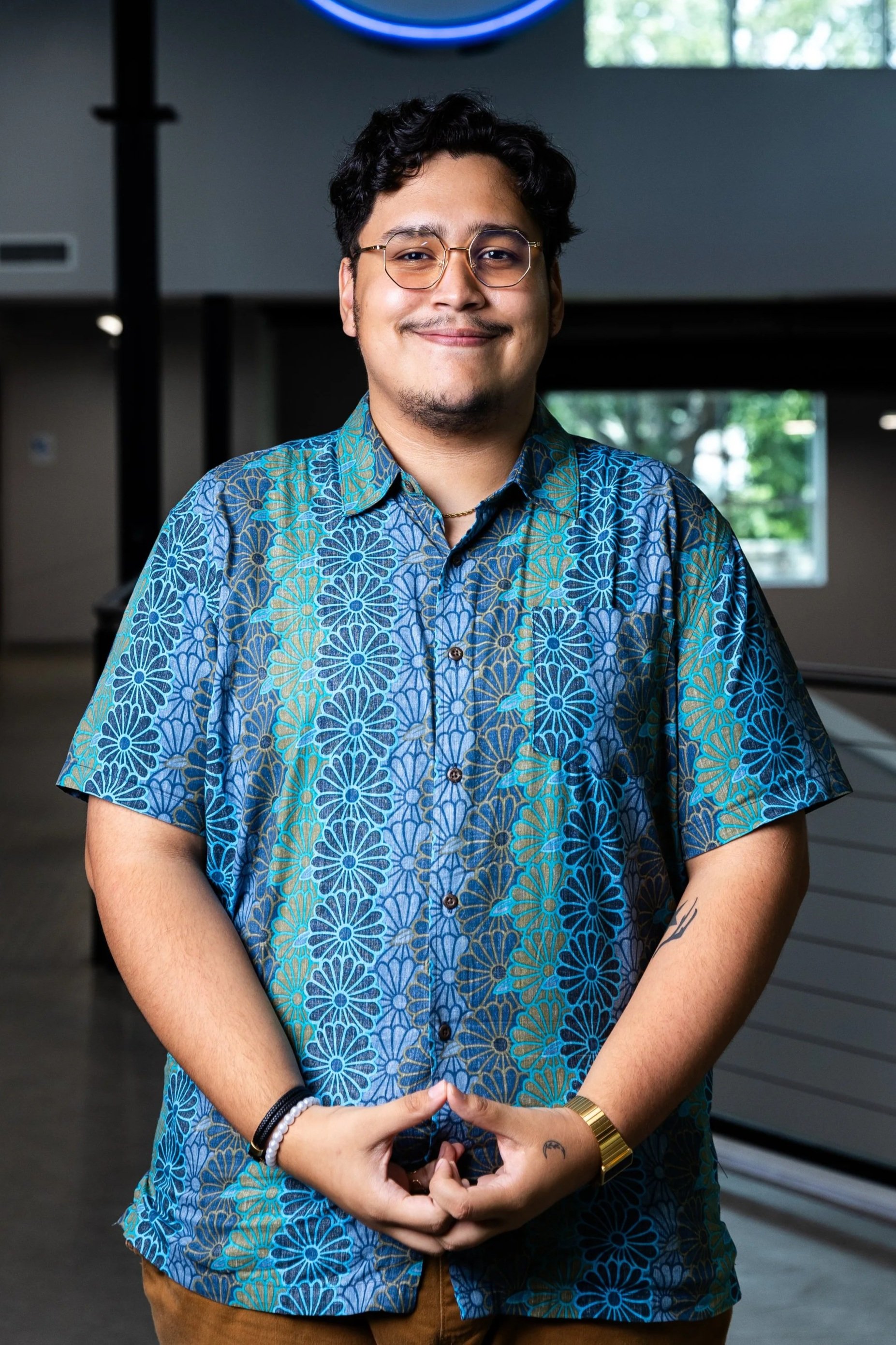 A young man with glasses and dark hair smiling, wearing a colorful floral shirt, standing indoors with a window and a neon sign in the background.