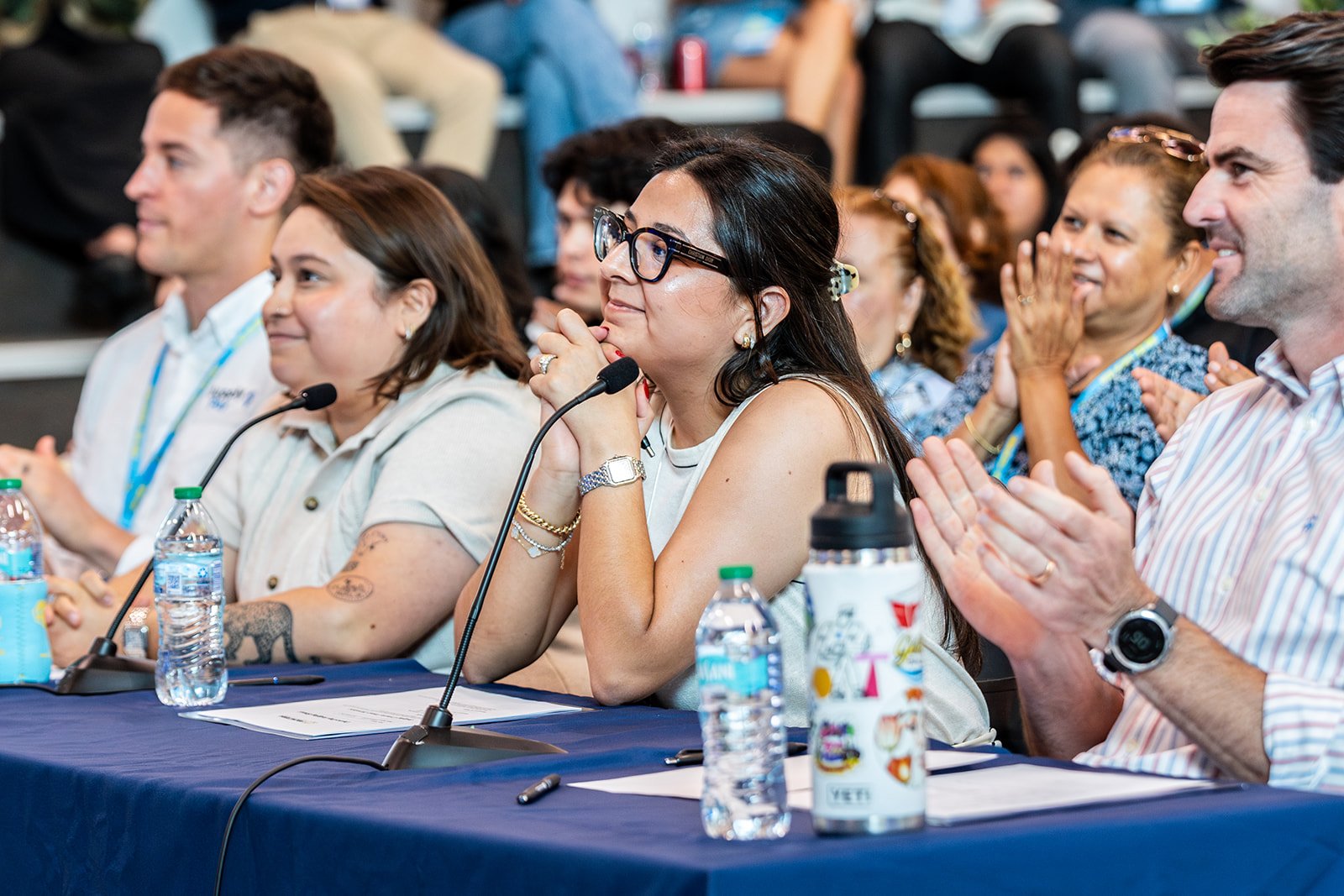 People attending a conference or seminar, sitting at a table with microphones, water bottles, and notes, clapping and listening to a speaker.