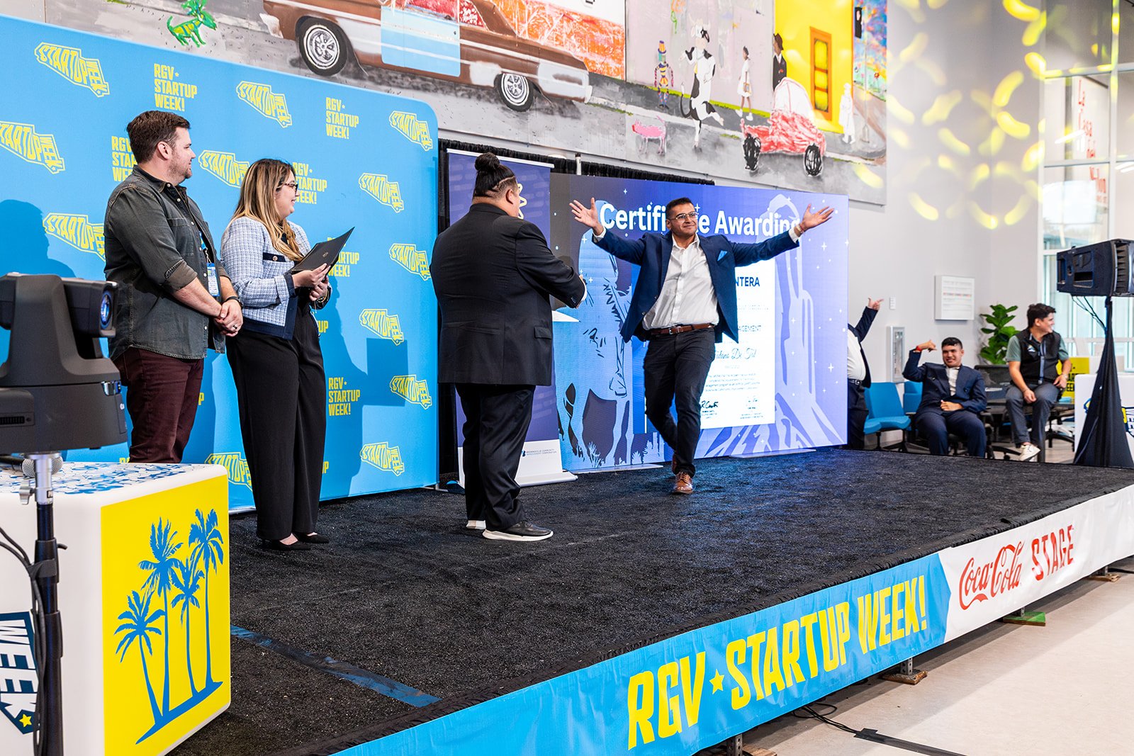 People on stage at an award ceremony during RGW Startup Week, with a large screen displaying 'Certificate Awarding' and a man with arms raised celebrating. The stage has a blue backdrop with 'RGW Startup Week' logos, and the area outside the stage has seated people and colorful artwork on the wall.