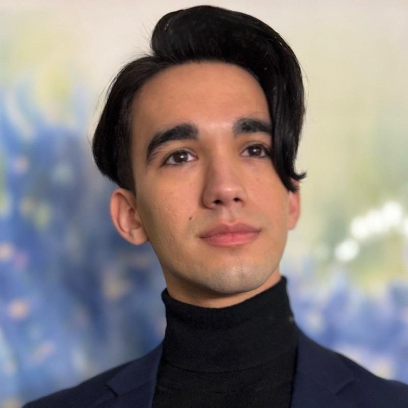 Portrait of a young man with dark hair, wearing a black turtleneck and blazer, in front of a blurred colorful background.